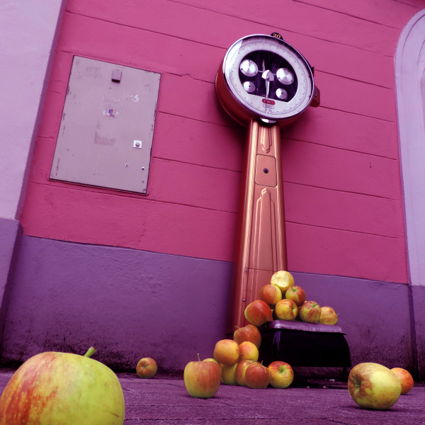 <p>Das Leben hat Gewicht - Projektbild Waage mit herunterfallenden Äpfeln in Pink vor einer Wand/Eingangstüre</p>